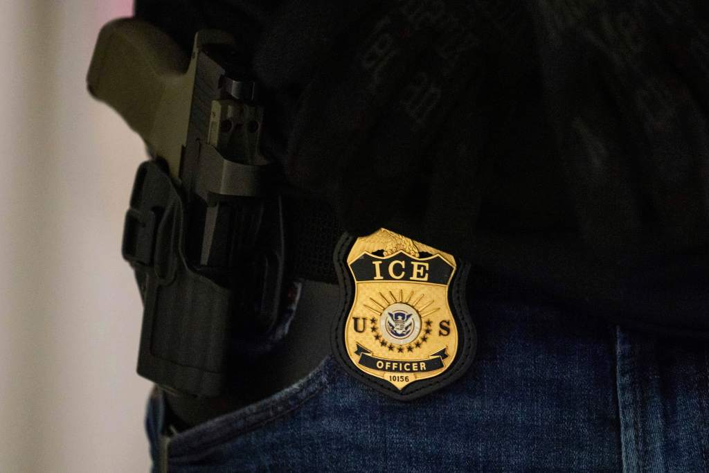 Close-up of an ICE officer’s badge and holstered firearm attached to a belt, partially visible against dark clothing and blue jeans.