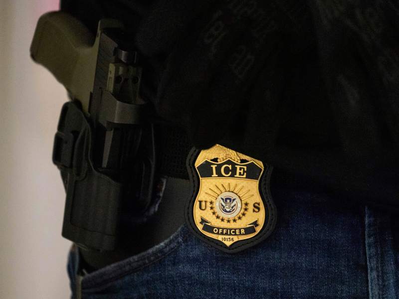 Close-up of an ICE officer’s badge and holstered firearm attached to a belt, partially visible against dark clothing and blue jeans.