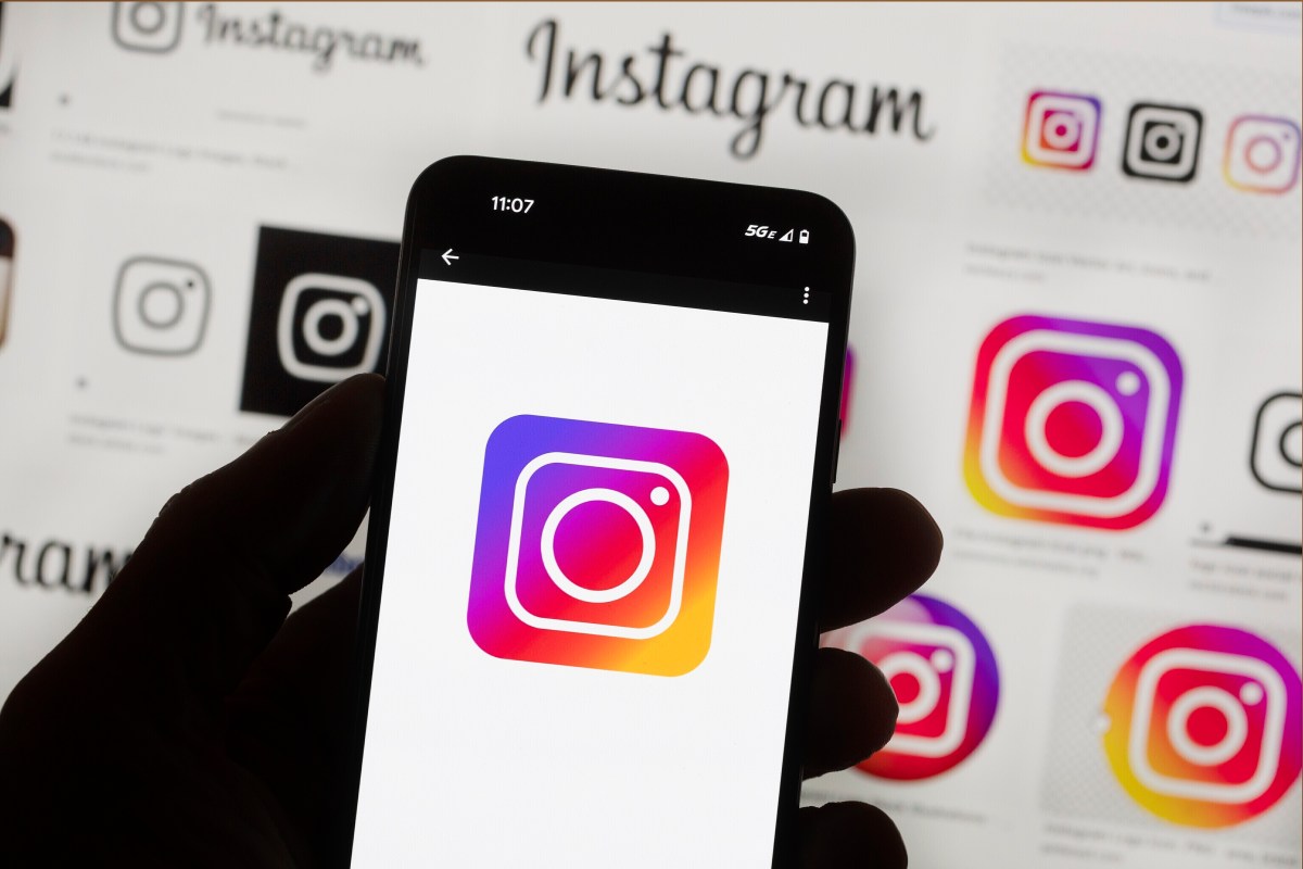 A hand holds a smartphone displaying the Instagram logo, with various versions of the Instagram logo shown on a screen in the background.