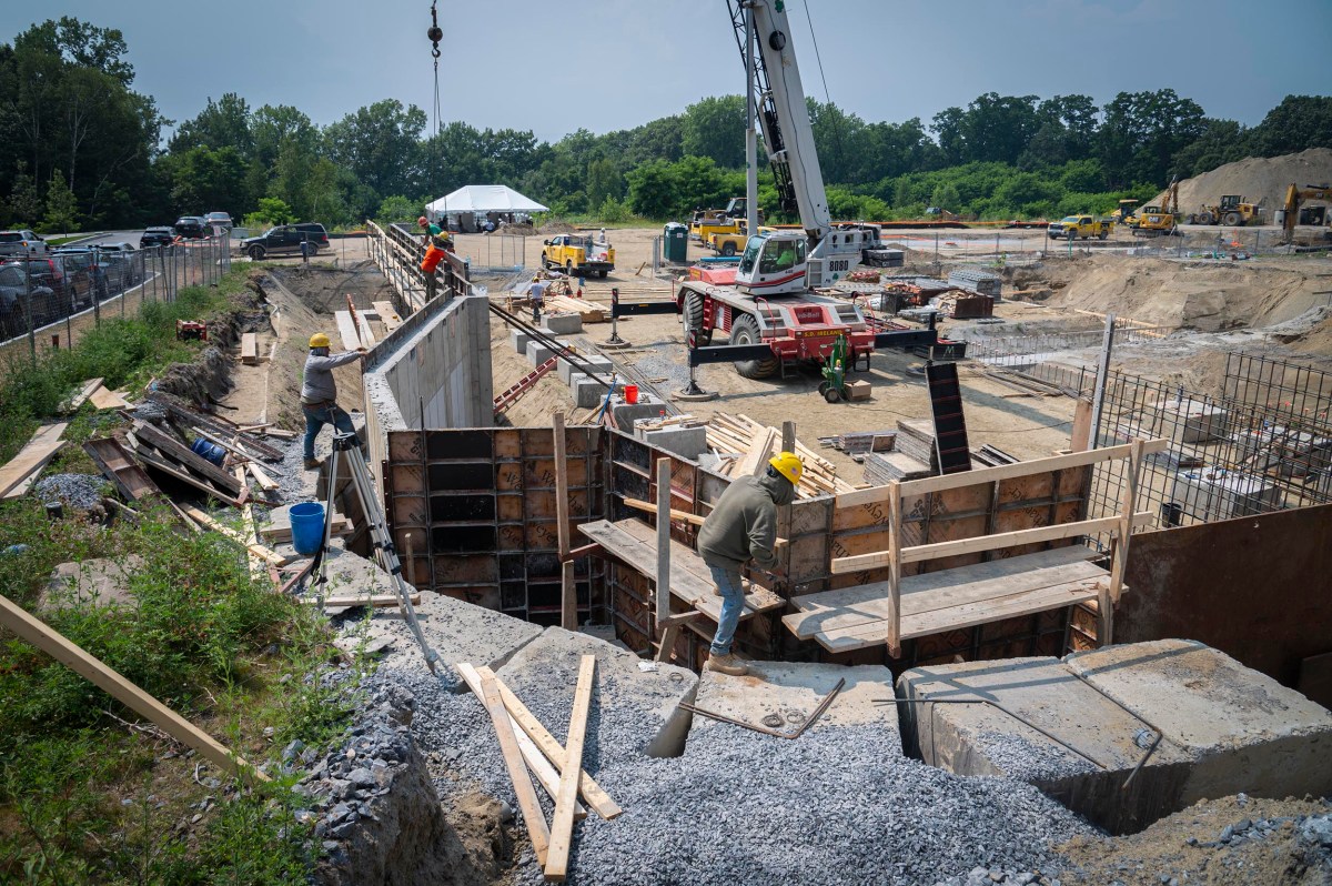 Construction workers wearing safety gear work on a building foundation at an active construction site with cranes, machinery, and materials visible.