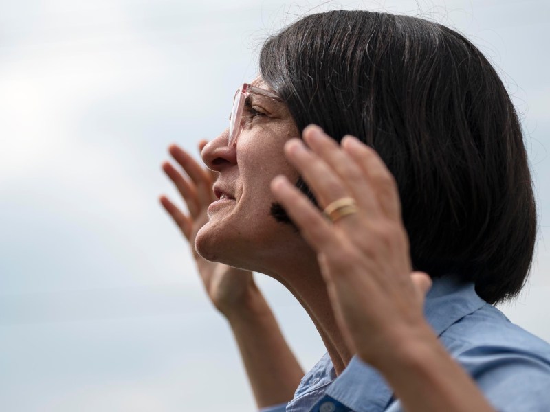 Person with short dark hair and glasses raises hands while speaking outdoors, wearing a blue shirt and a gold ring, against a light sky background.