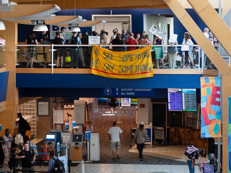 A group of people protest on an airport balcony with signs and a yellow banner reading “If you see something, we say something” above a security checkpoint area.