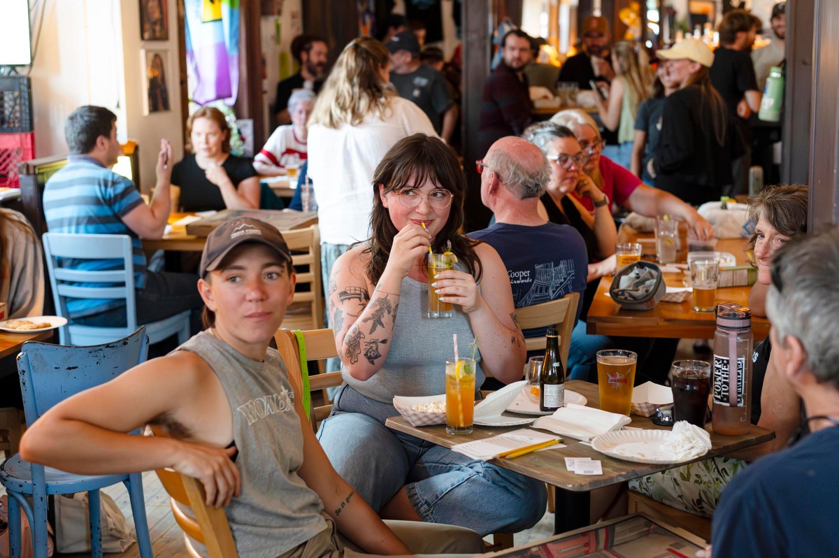A group of people sits at tables in a busy restaurant or café, eating, drinking, and socializing. One person sips a drink and looks at the camera.