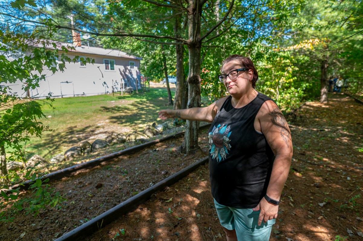 A woman wearing glasses and a sleeveless shirt stands outdoors, pointing toward a house in the background surrounded by trees and greenery.