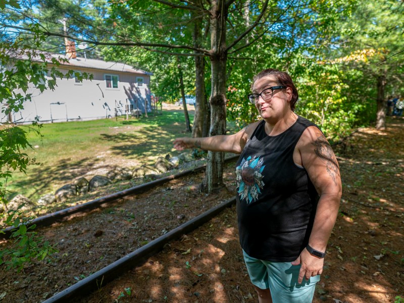 A woman wearing glasses and a sleeveless shirt stands outdoors, pointing toward a house in the background surrounded by trees and greenery.
