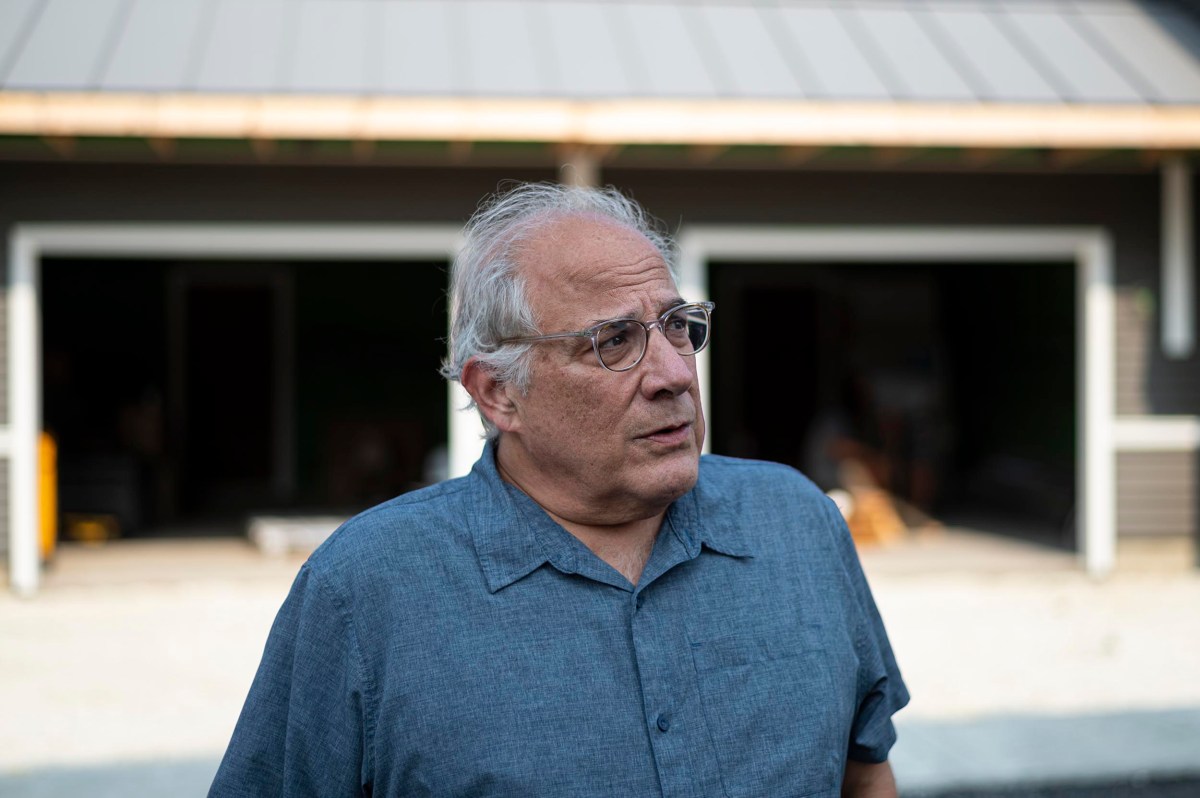 An older man with gray hair and glasses stands outside in front of a building with two open garage doors, wearing a short-sleeve button-up shirt.