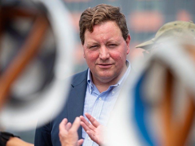 A man in a suit stands outdoors, facing the camera, with blurred objects and people in the foreground.