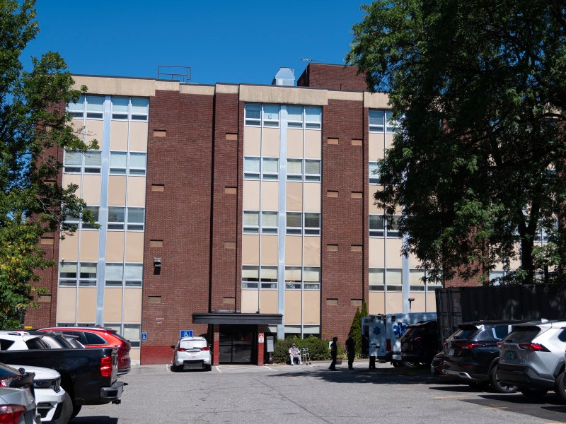 A five-story brick building with vertical beige panels, surrounded by parked cars and trees, under a clear blue sky.