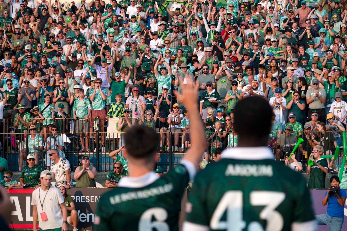 Two soccer players in green jerseys face a large, cheering crowd in the stands, many wearing green and raising their hands in support.