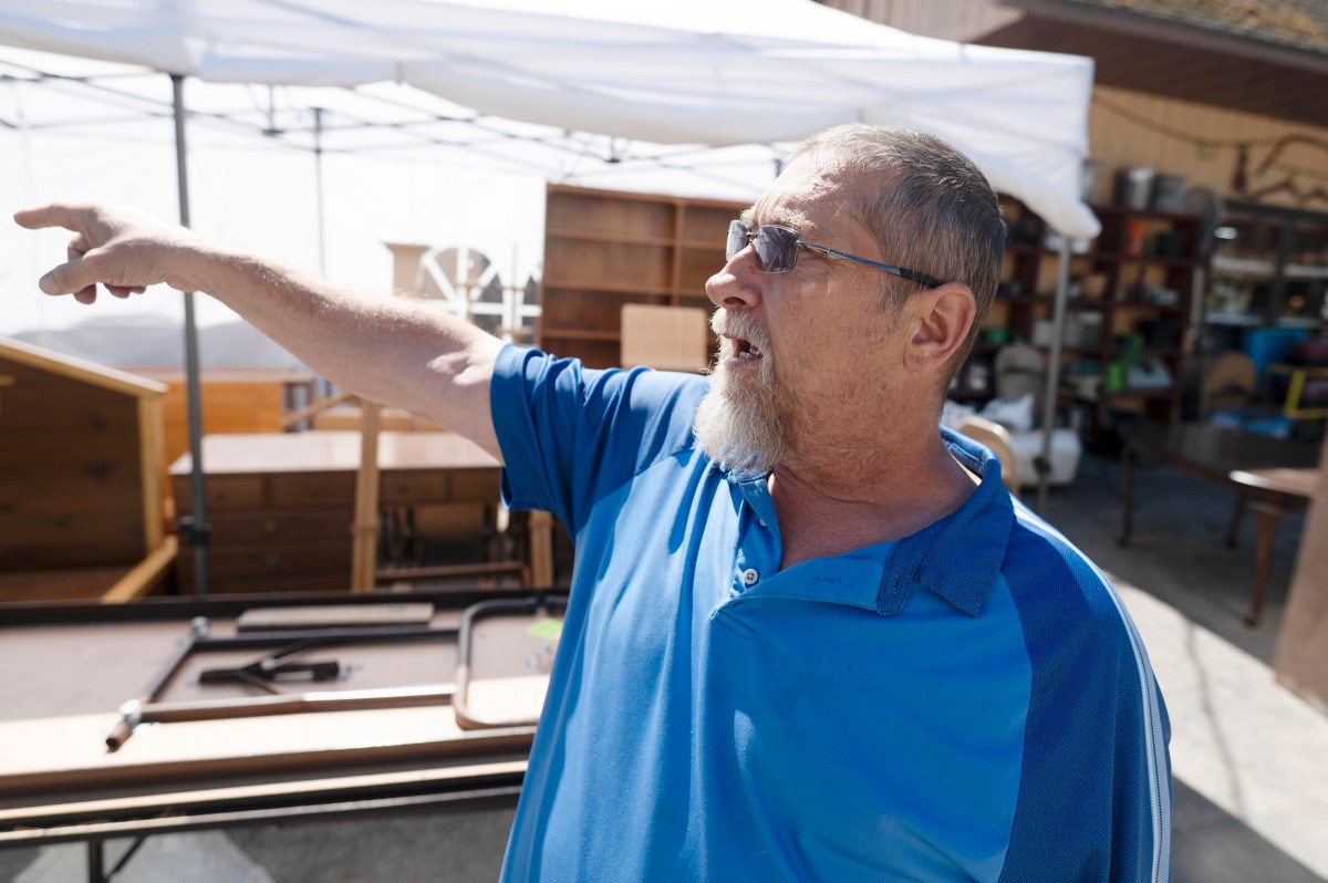 A man in a blue shirt and sunglasses stands outdoors near wooden furniture, pointing to something off-camera.