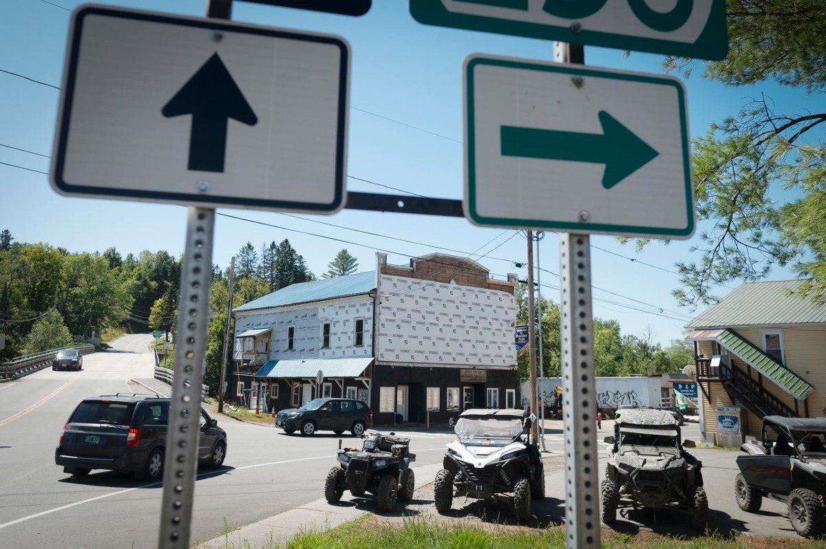 Two road signs, one with a straight arrow and one with a right arrow, stand at an intersection with cars and ATVs parked near buildings under a clear sky.