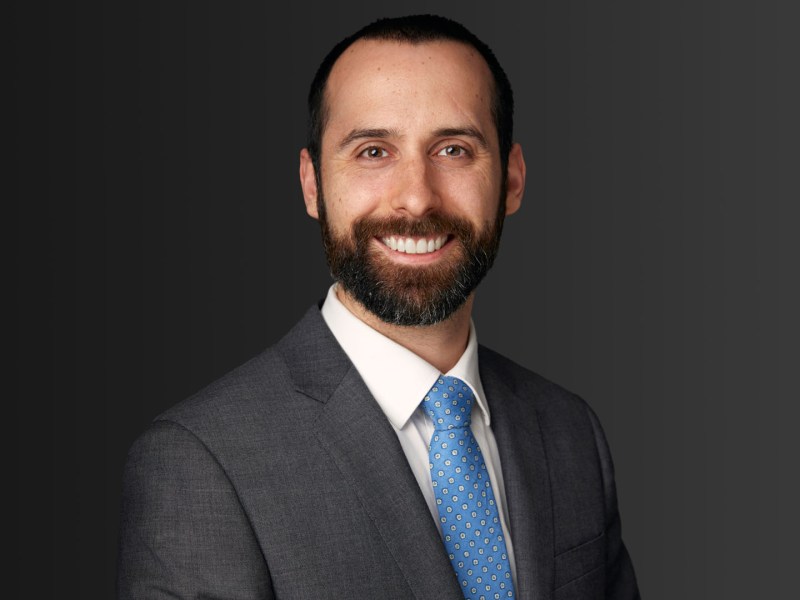 A man with a beard and short dark hair, wearing a gray suit, white shirt, and blue dotted tie, smiles in front of a dark gradient background.