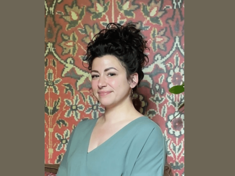 A woman with curly dark hair wearing a green top sits in front of a red and beige patterned background, looking at the camera and smiling slightly.