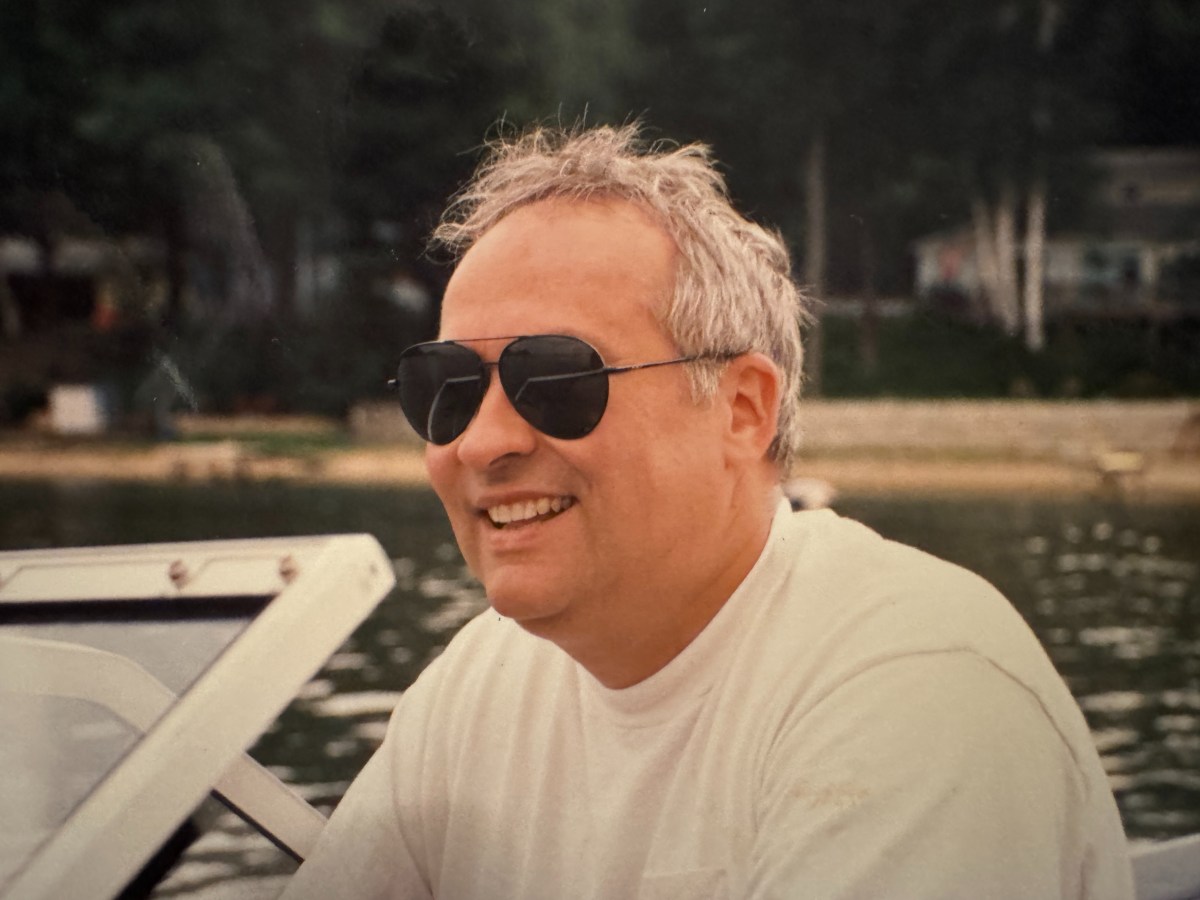 A man wearing sunglasses and a white t-shirt smiles while sitting on a boat, with water and trees visible in the background.
