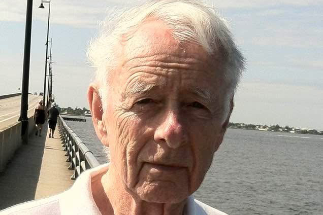 A man stands by a waterfront walkway on a sunny day, with a bridge and water in the background.