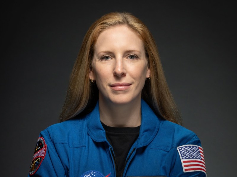 A woman in a blue NASA flight suit with mission patches and an American flag patch poses against a dark background.