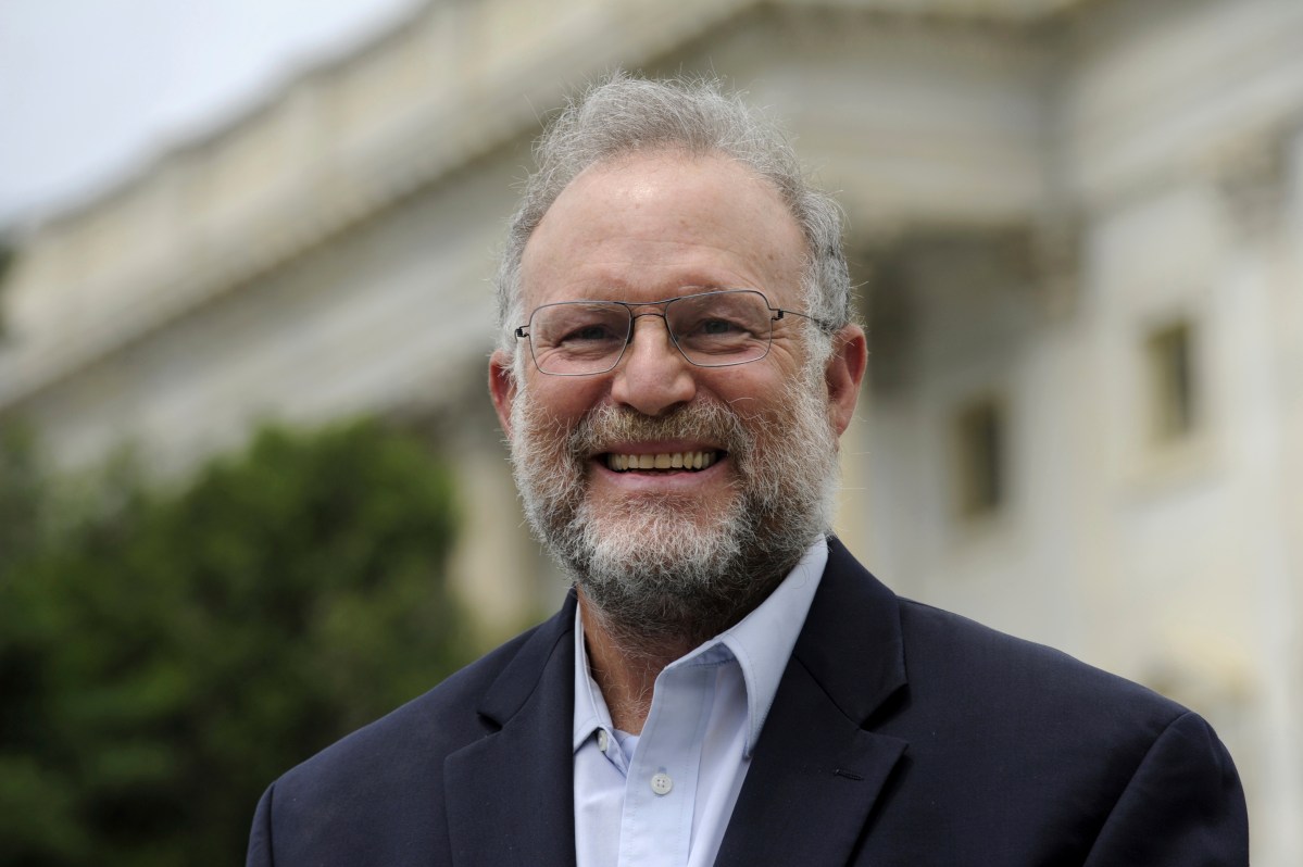 A man with gray hair, a beard, and glasses smiles while wearing a dark suit jacket and light blue shirt, standing outdoors in front of a blurred building.
