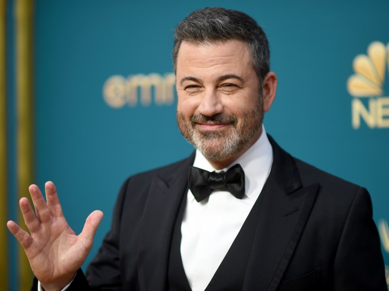 A man in a black tuxedo waves and smiles while standing in front of a teal background with gold and NBC logos.