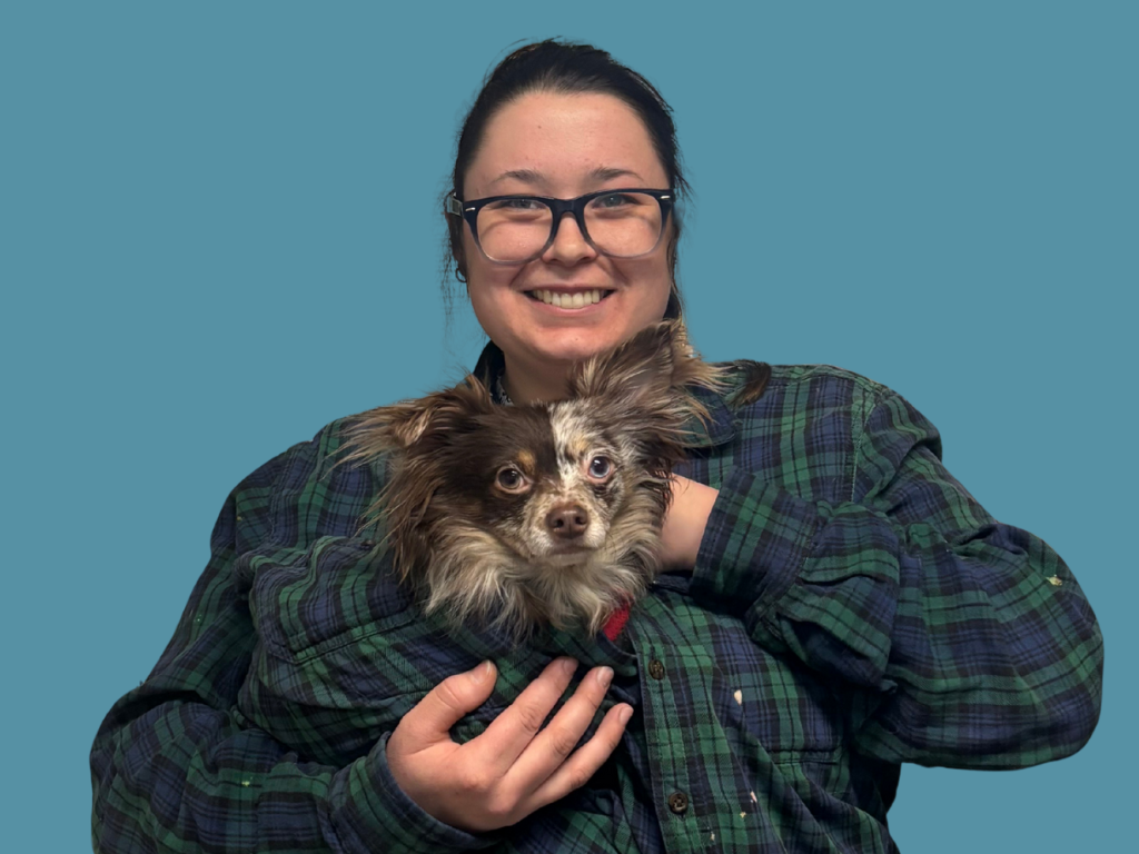 A person wearing glasses and a green plaid shirt smiles while holding a small brown and white dog against a plain blue background.
