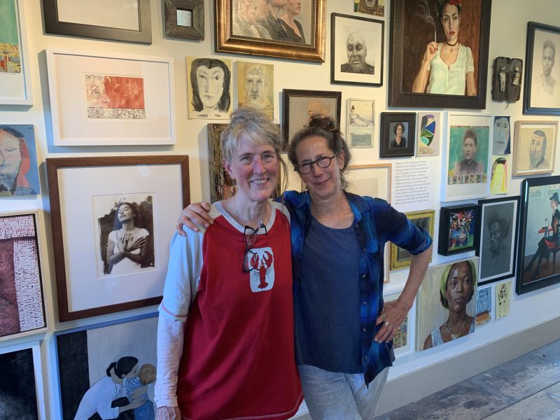 Two women stand smiling with arms around each other in front of a wall filled with diverse framed artwork and portraits in a gallery setting.
