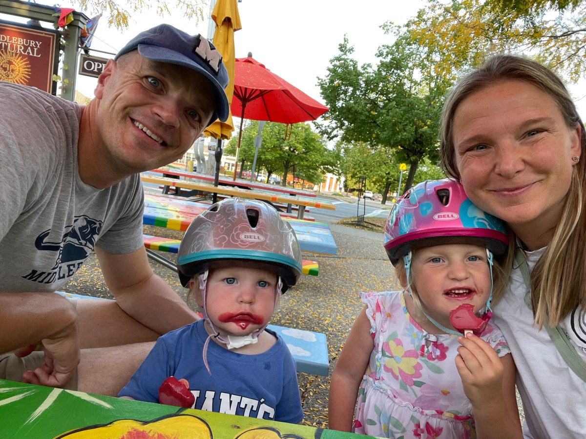 Two adults and two young children wearing helmets sit at a colorful outdoor table, eating watermelon slices. Trees and umbrellas are visible in the background.