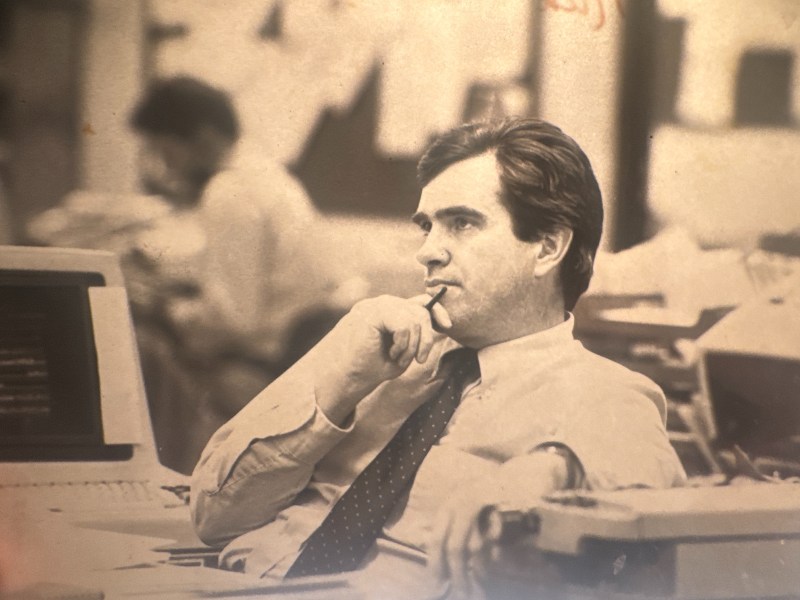 A man in a dress shirt and tie sits at an office desk, holding a pen to his mouth, appearing thoughtful. Computer equipment and blurred coworkers are visible in the background.