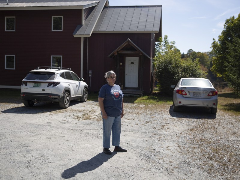 A person stands in a gravel driveway between two parked cars in front of a red building on a sunny day.