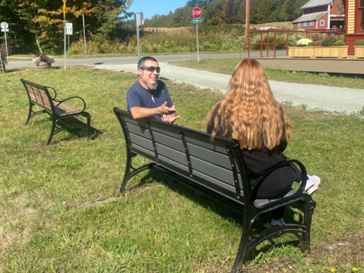Two people sit on a bench in a grassy area near a road, talking. There are additional benches, a stop sign, and a red building visible in the background.