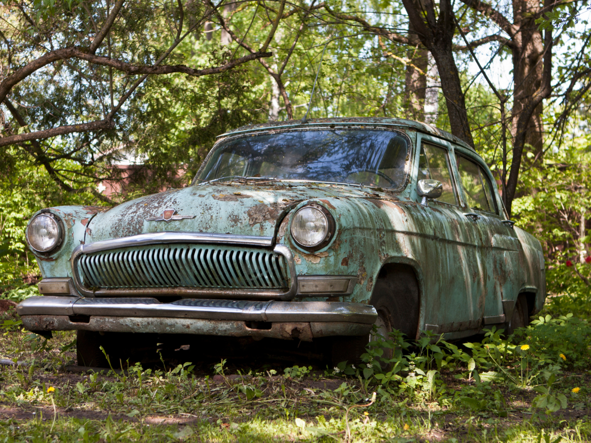 An old, rusted car with faded teal paint sits abandoned in a grassy, wooded area under tree shade.