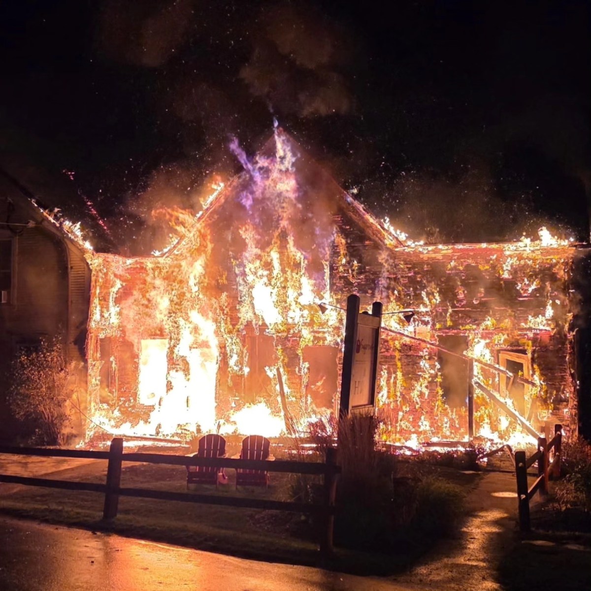 A house is engulfed in flames at night, with fire covering the roof and walls while smoke rises into the dark sky.