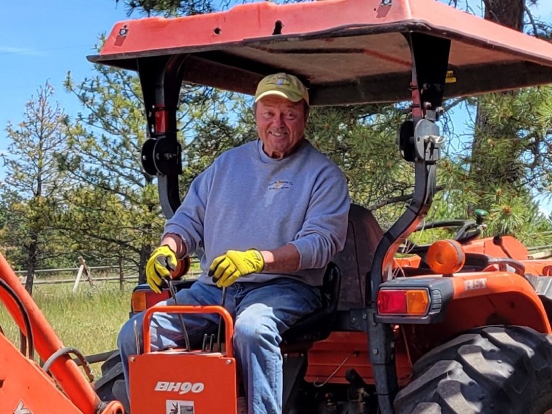 Man wearing a gray sweatshirt, yellow gloves, and cap sits on an orange tractor outside, surrounded by trees and grass.