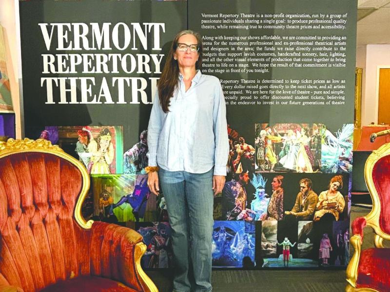 A woman stands in front of a Vermont Repertory Theatre display, flanked by ornate chairs, with images and text about the theatre in the background.