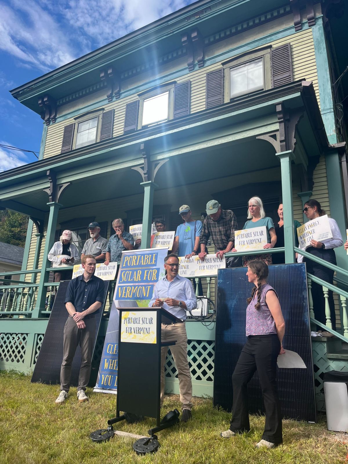 A group of people stand on the porch of a large house holding signs that read "Portable Solar for Vermont" during an outdoor press event.