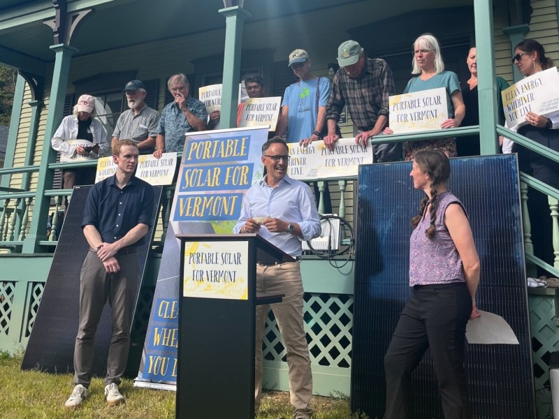 A group of people stand on a porch holding signs that read "Portable Solar for Vermont" during an outdoor event or press conference.