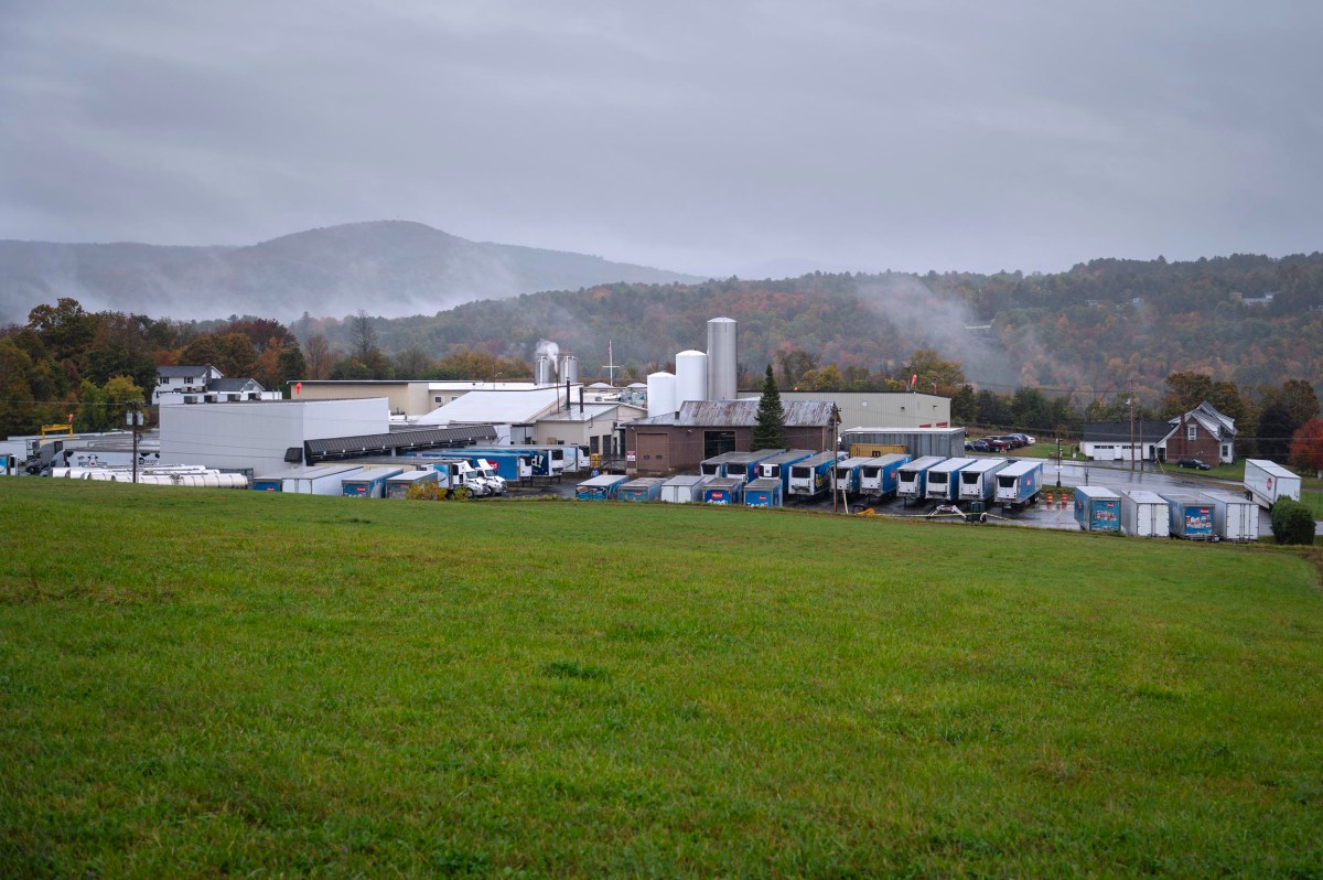 Industrial facility with multiple buildings, white storage tanks, and loading docks, situated on a grassy field with misty hills and autumn trees in the background.