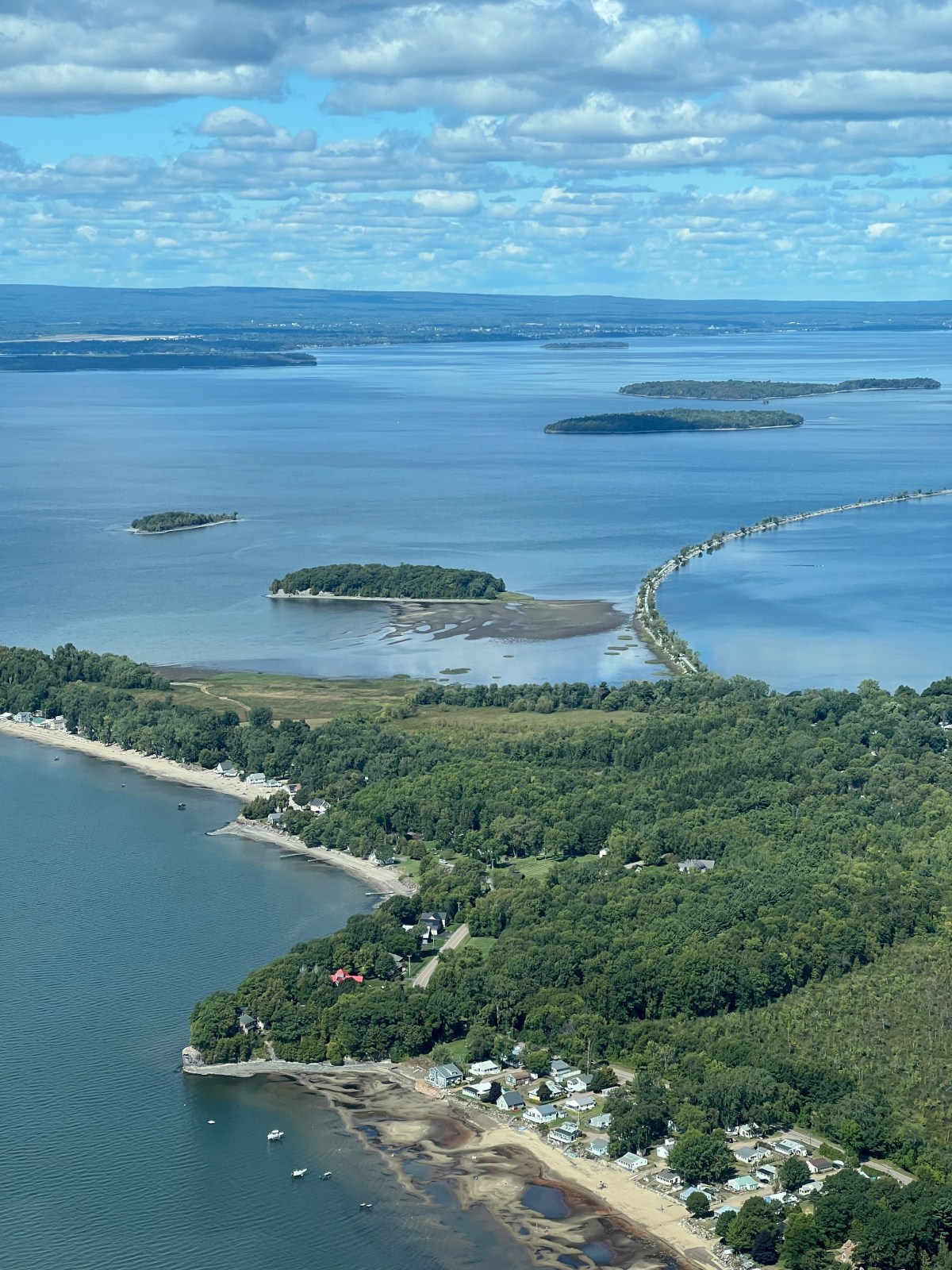 Aerial view of a peninsula with houses, dense green trees, sandy shores, and a narrow road extending across the water to several small islands.
