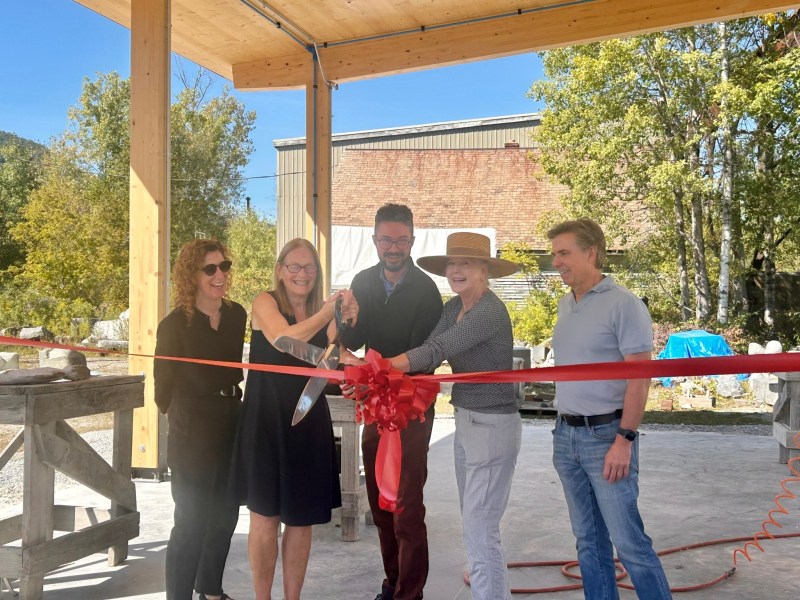 Five people stand together cutting a red ribbon with large scissors at an outdoor event under a wooden structure, with buildings and trees in the background.