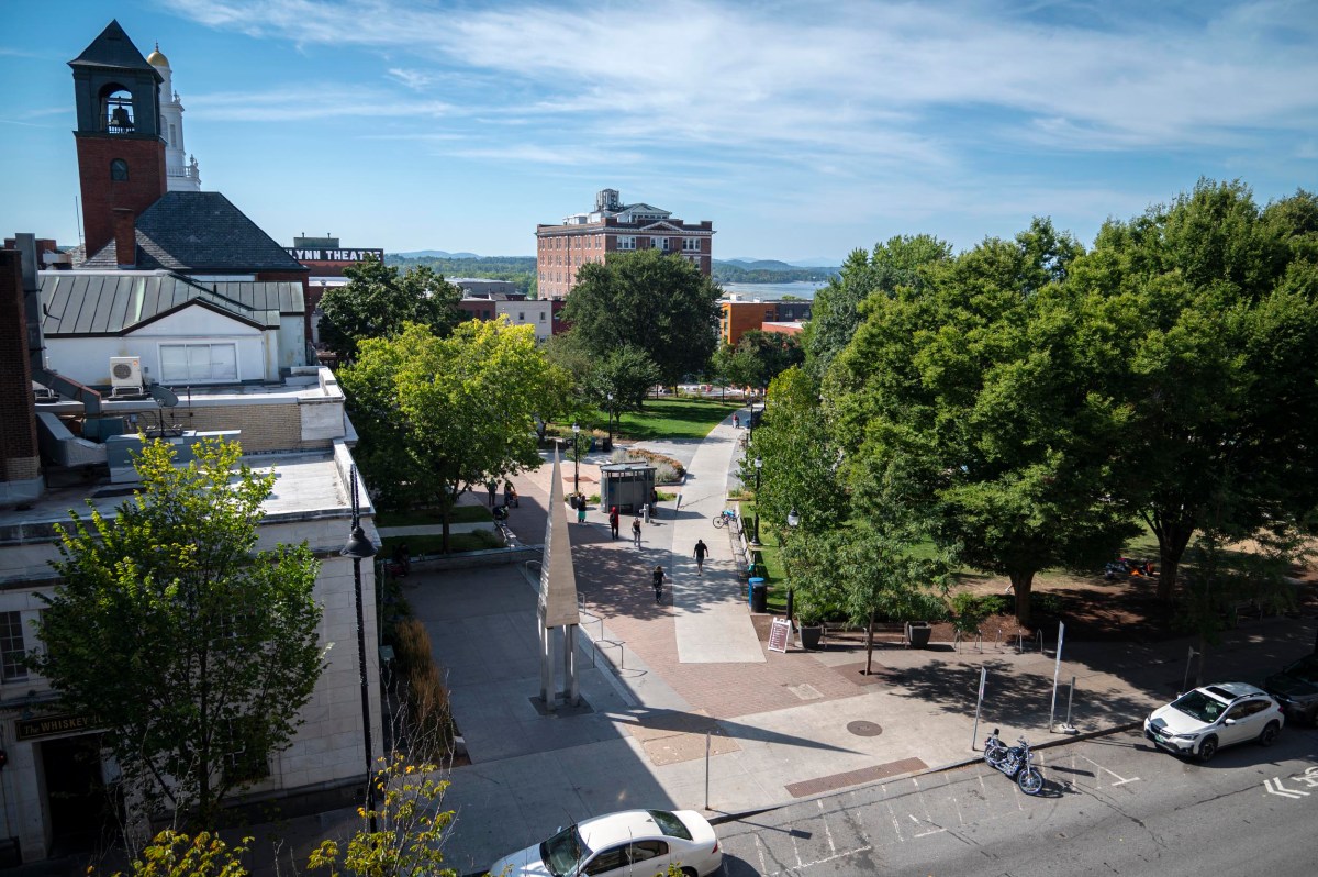 A city square with trees, pedestrians, parked bicycles, and surrounding buildings under a blue sky with scattered clouds.