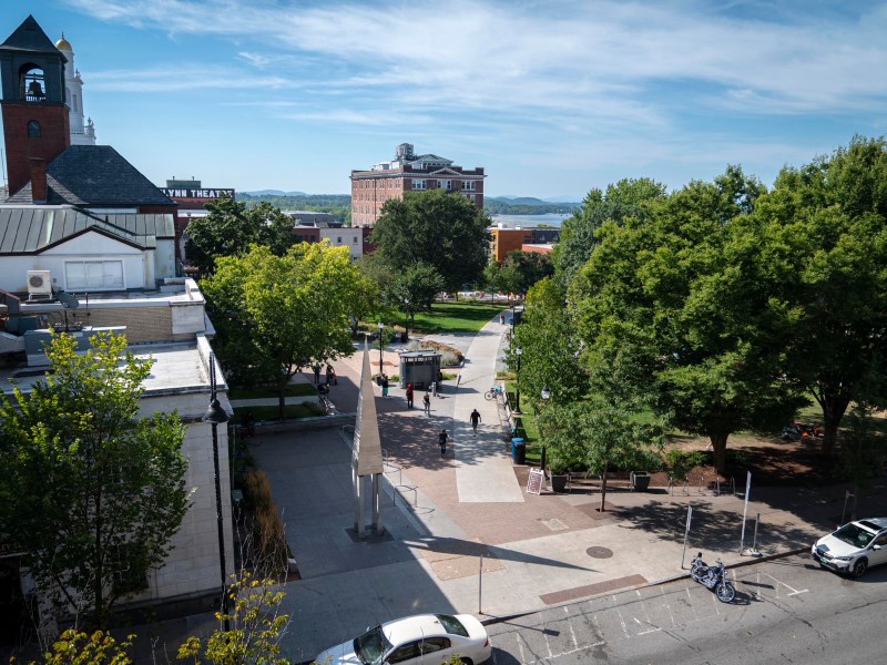 A city square with trees, pedestrians, parked bicycles, and surrounding buildings under a blue sky with scattered clouds.