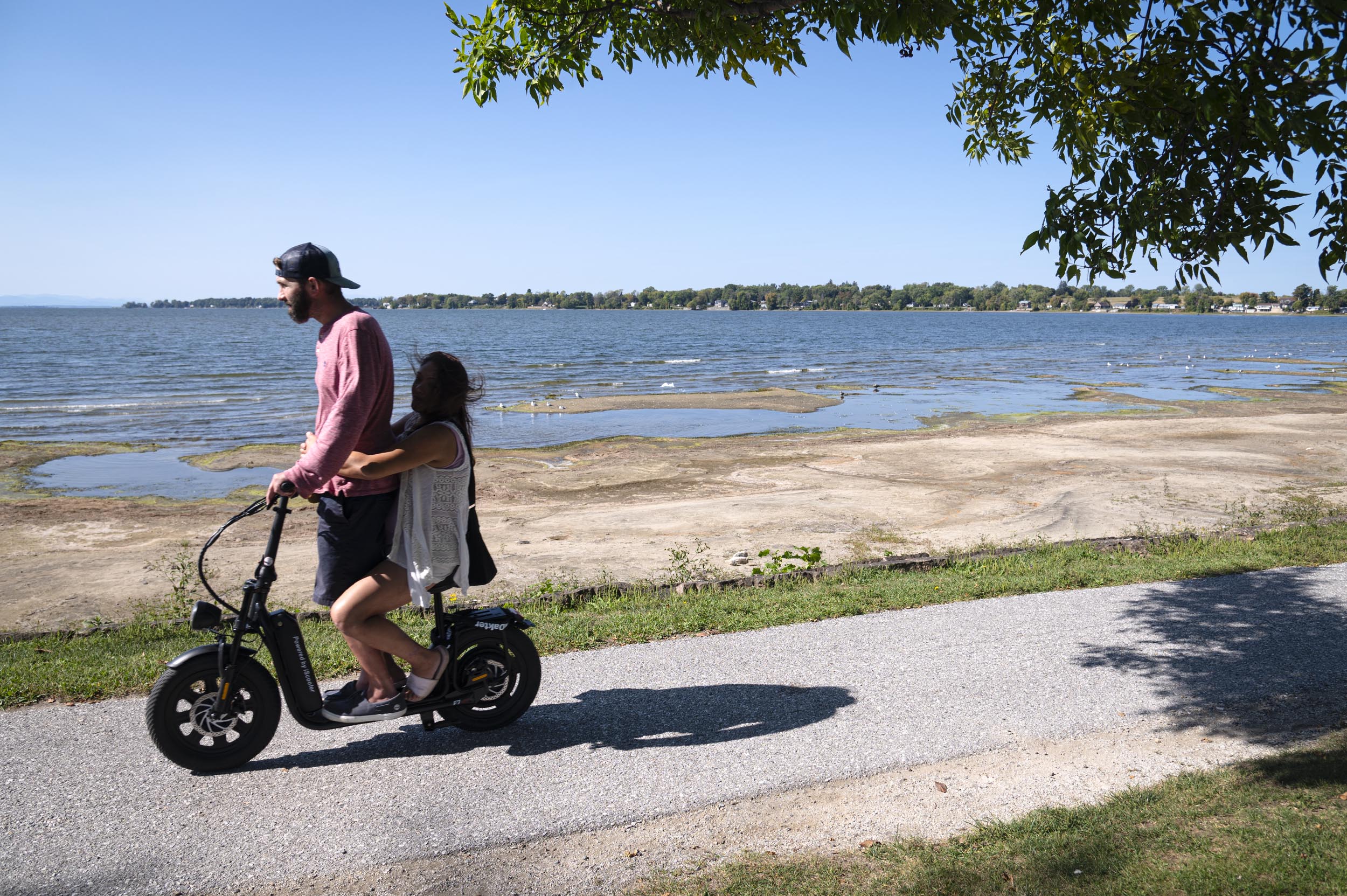 A man and a woman ride together on an electric scooter along a paved path by a lake, with trees and distant shoreline visible under a clear sky.