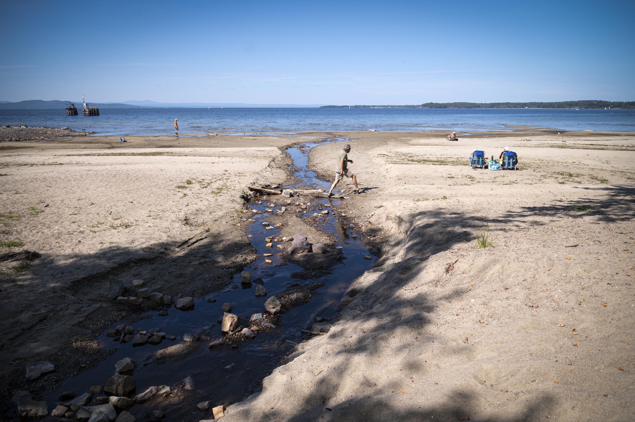 A person walks across a shallow stream on a sandy beach with a few people sitting and standing in the distance under a clear blue sky.