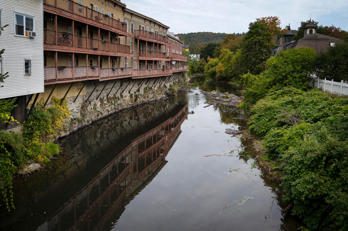 A narrow river runs between a building with balconies on stilts and dense green vegetation, with trees and a hill visible in the background.