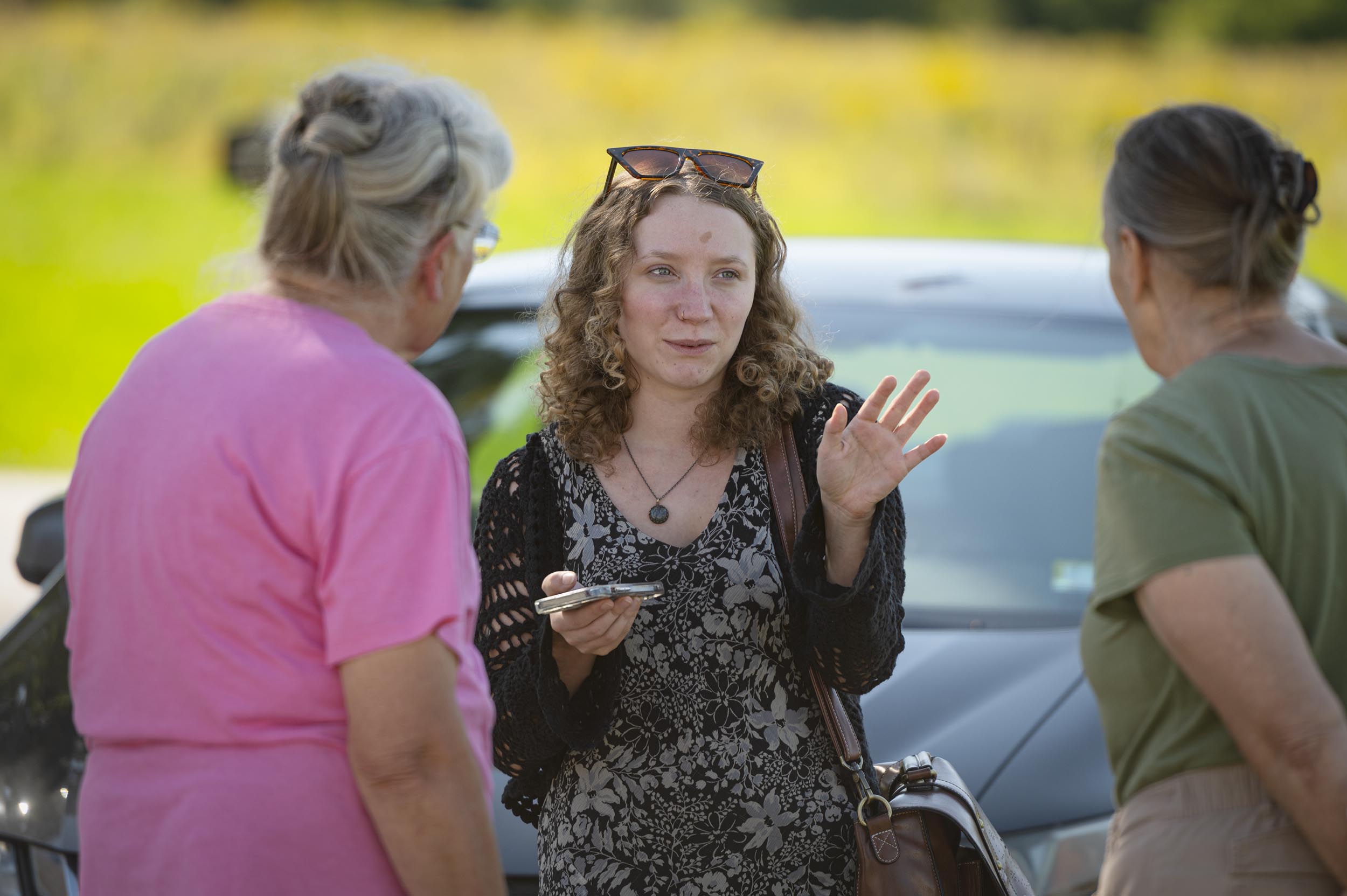 Three women stand and talk outdoors in front of a parked car; one woman gestures with her hand while holding a phone.