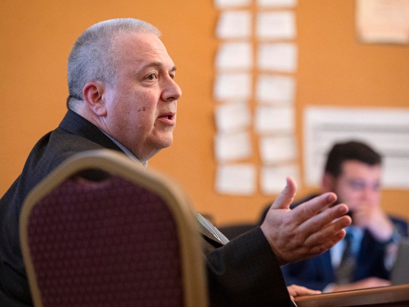 A man in a suit speaks while seated at a table in a meeting room; another person is visible in the background, blurred.