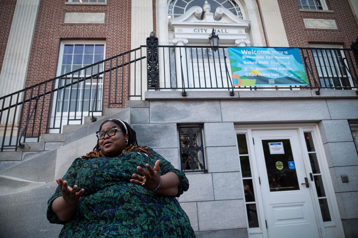 A person with glasses sits on steps outside a brick building with a "City Hall" sign above the entrance and a colorful banner on the railing.