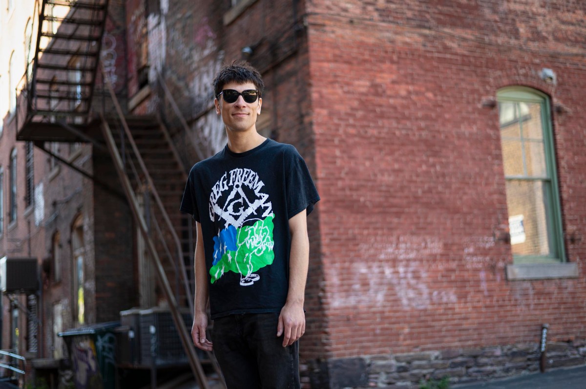 A person wearing sunglasses and a black graphic t-shirt stands outdoors in front of a brick building with a metal staircase.