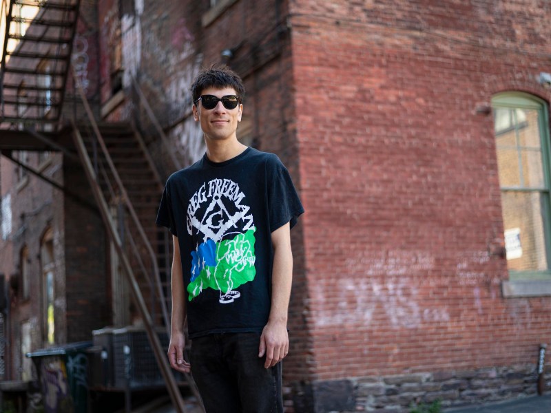 A person wearing sunglasses and a black graphic t-shirt stands outdoors in front of a brick building with a metal staircase.