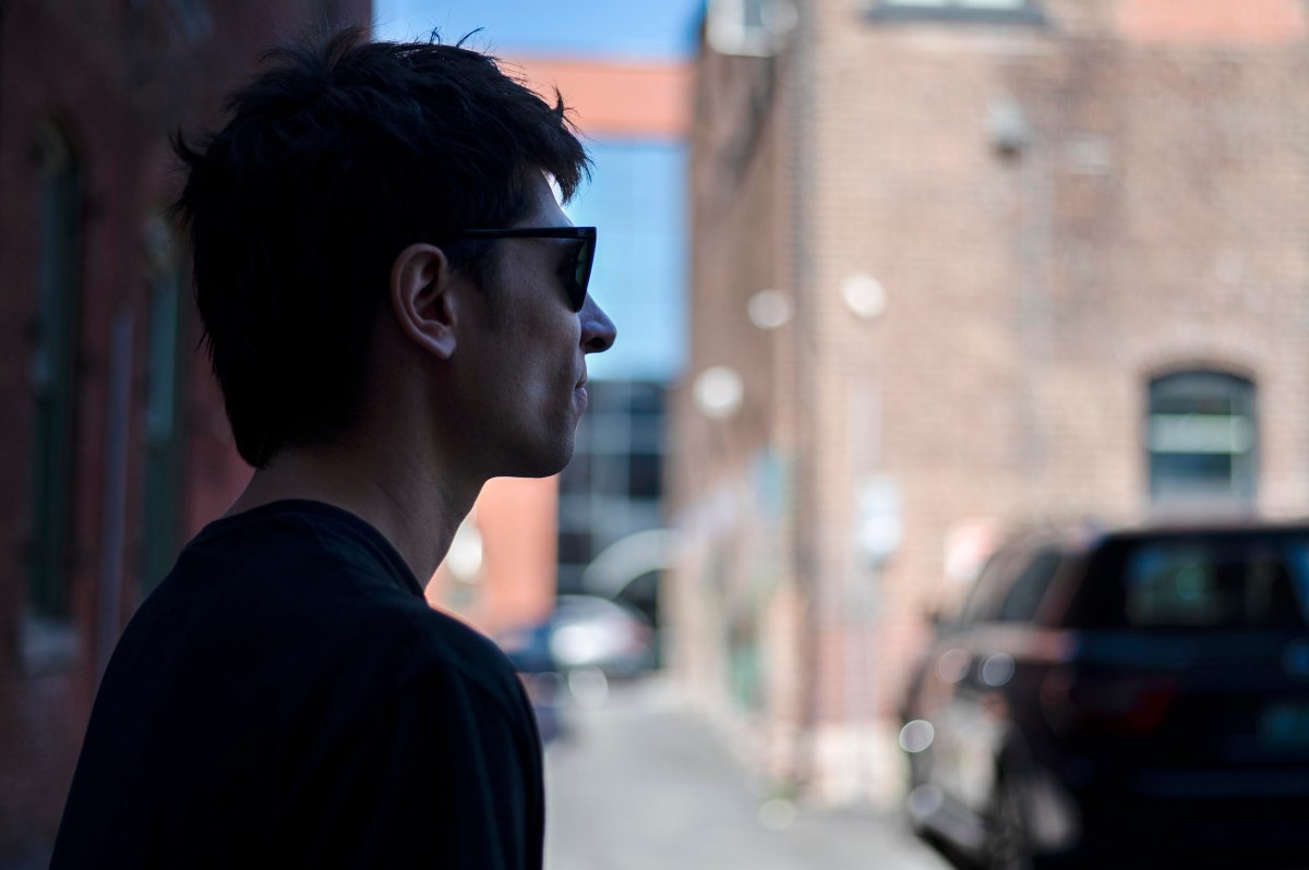 A person wearing sunglasses stands in profile in a narrow alleyway between brick buildings, with cars parked along the side and background out of focus.