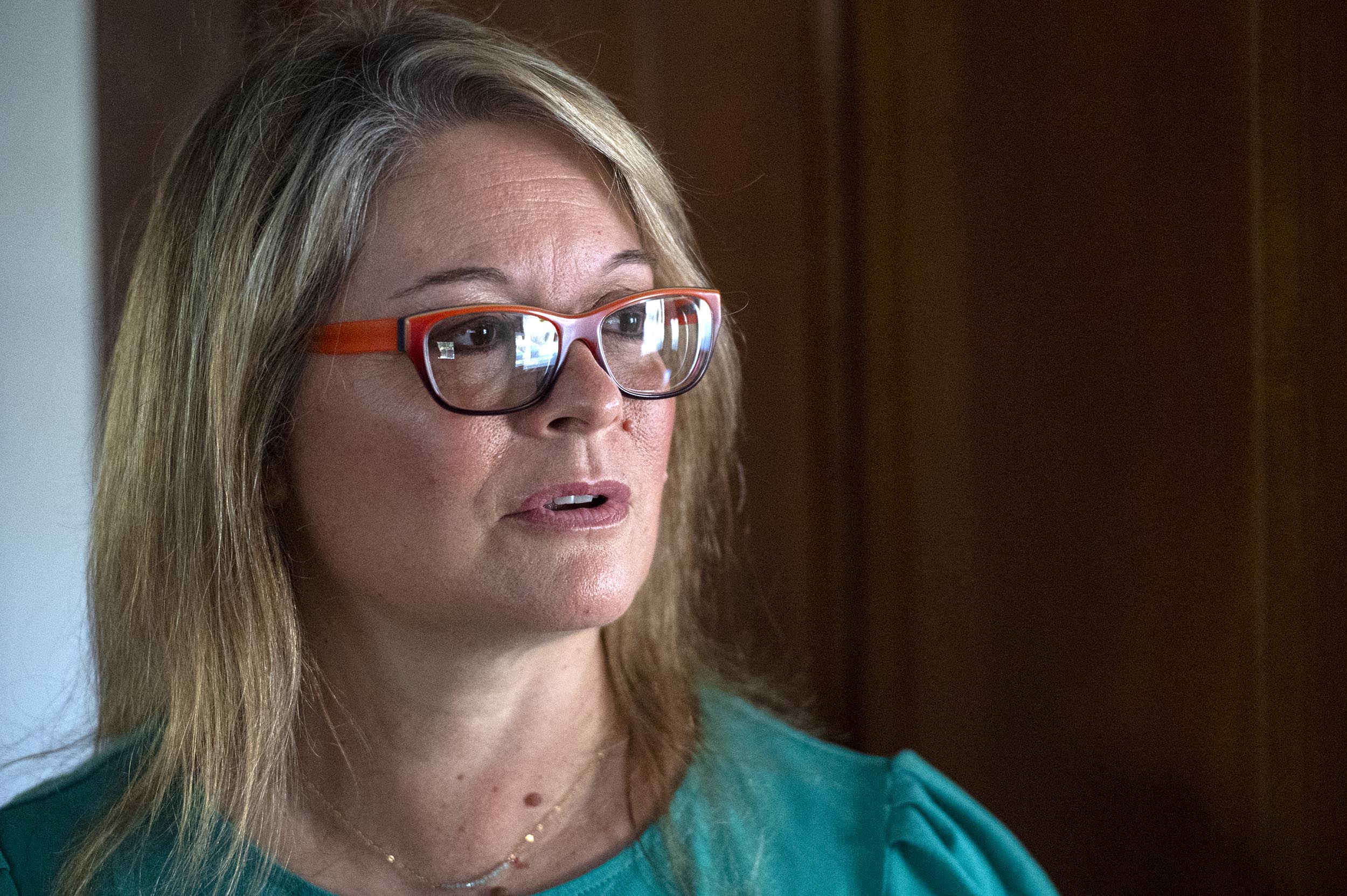 A woman with light brown hair and orange-framed glasses wearing a teal top stands indoors, looking slightly to the side.