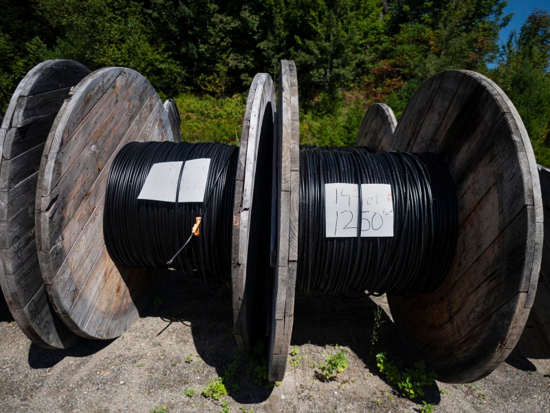 Large wooden spools of black cable are lined up outdoors on gravel, with handwritten labels displaying cable length and measurements attached to the spools.